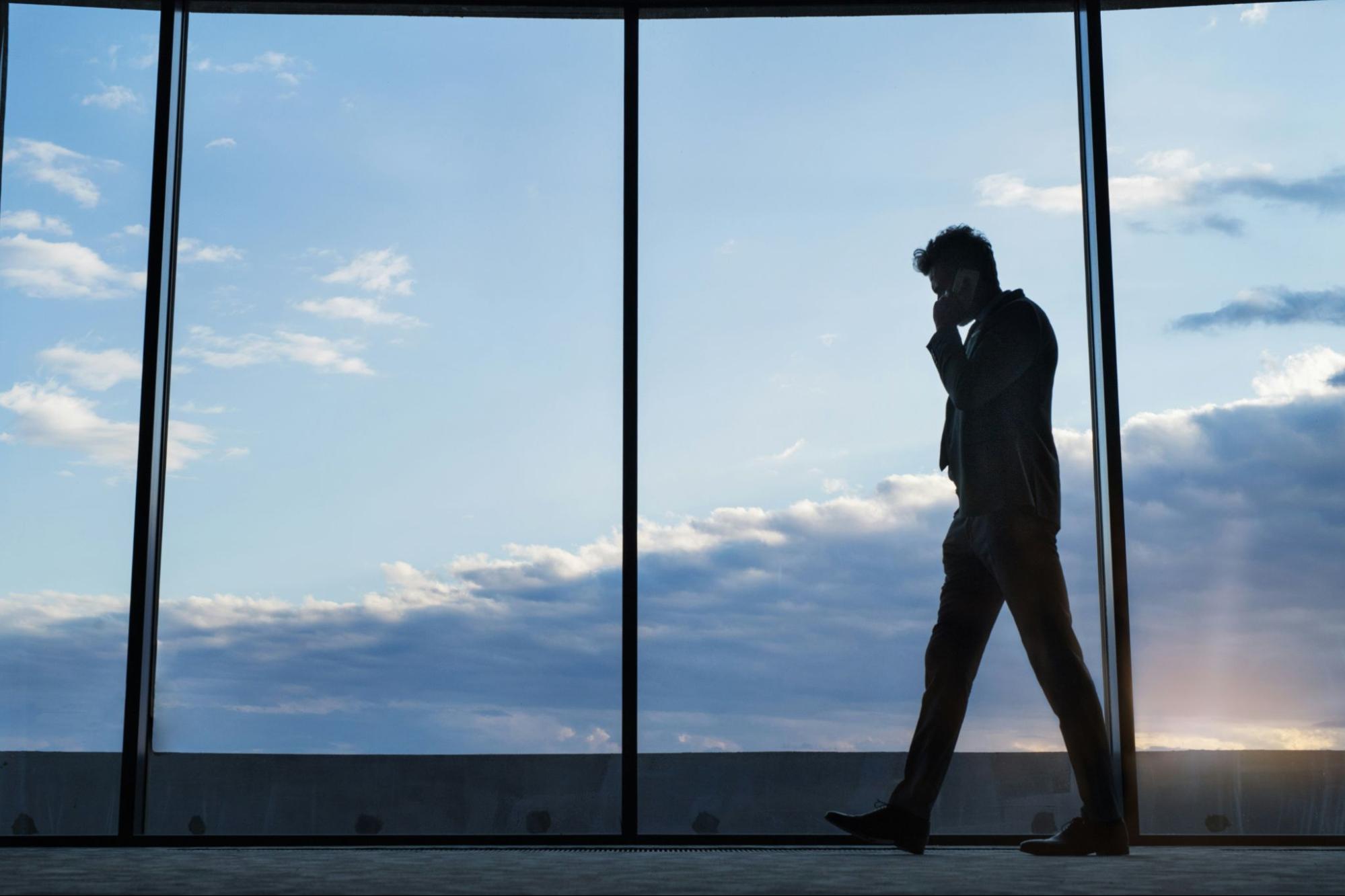 Business man walking in front of huge window in office building with sky and clouds visible through the window.