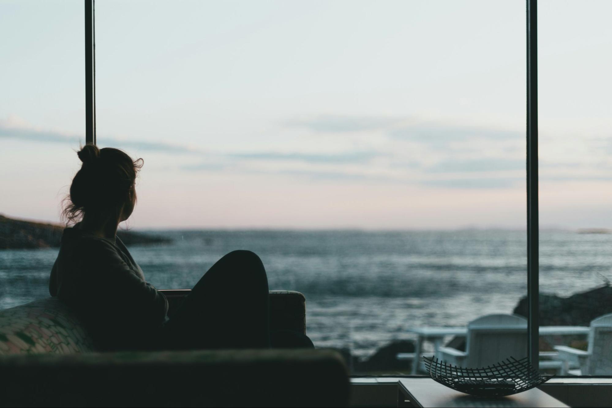 Woman sitting alone on couch in a dark room looking out of a winow to the ocean at sunset. 