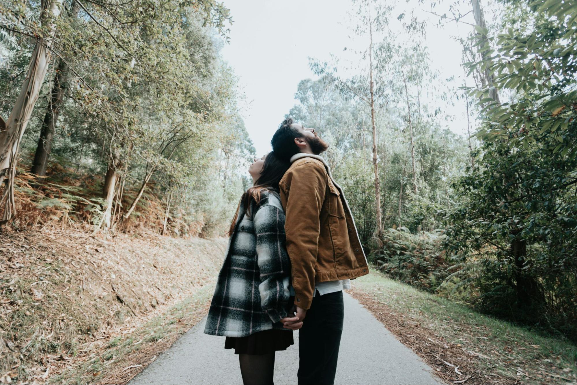 Couple standing back to back on a path through the woods, leaning on each other. 