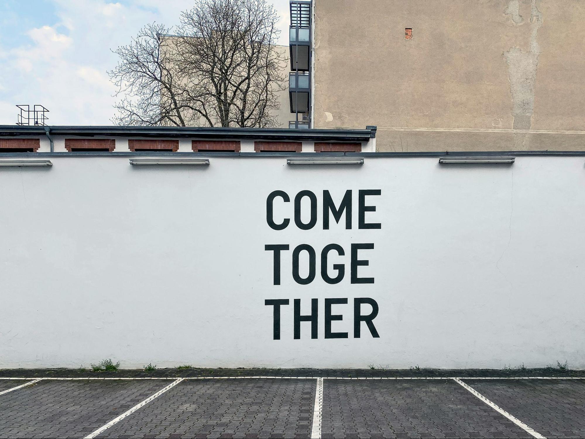 A white wall in a parking lot with the words "come together" painted on it in big letters. 
