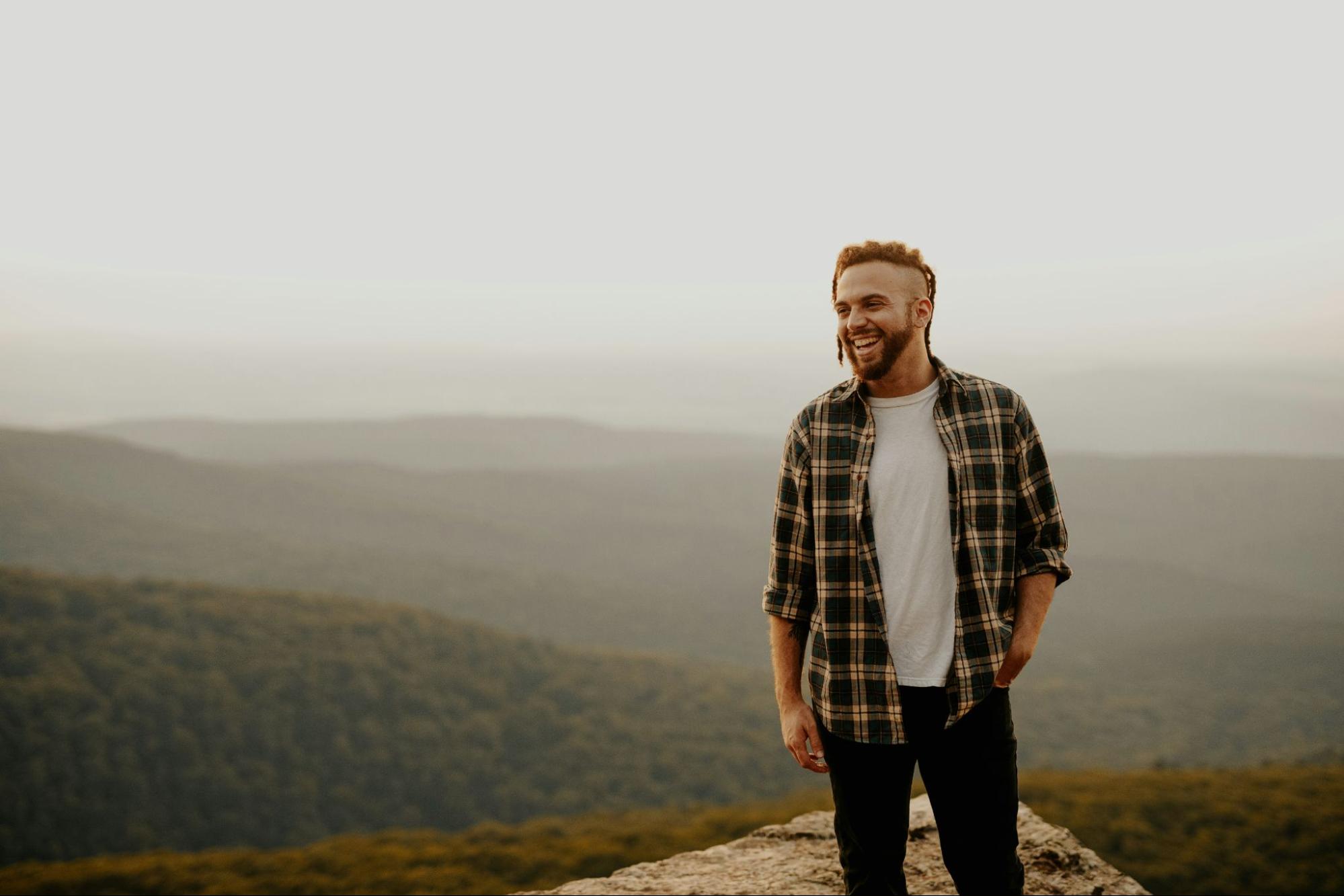 Man wearing a flannel and jeans standing a the top of a mountain relaxed and smiling. 
