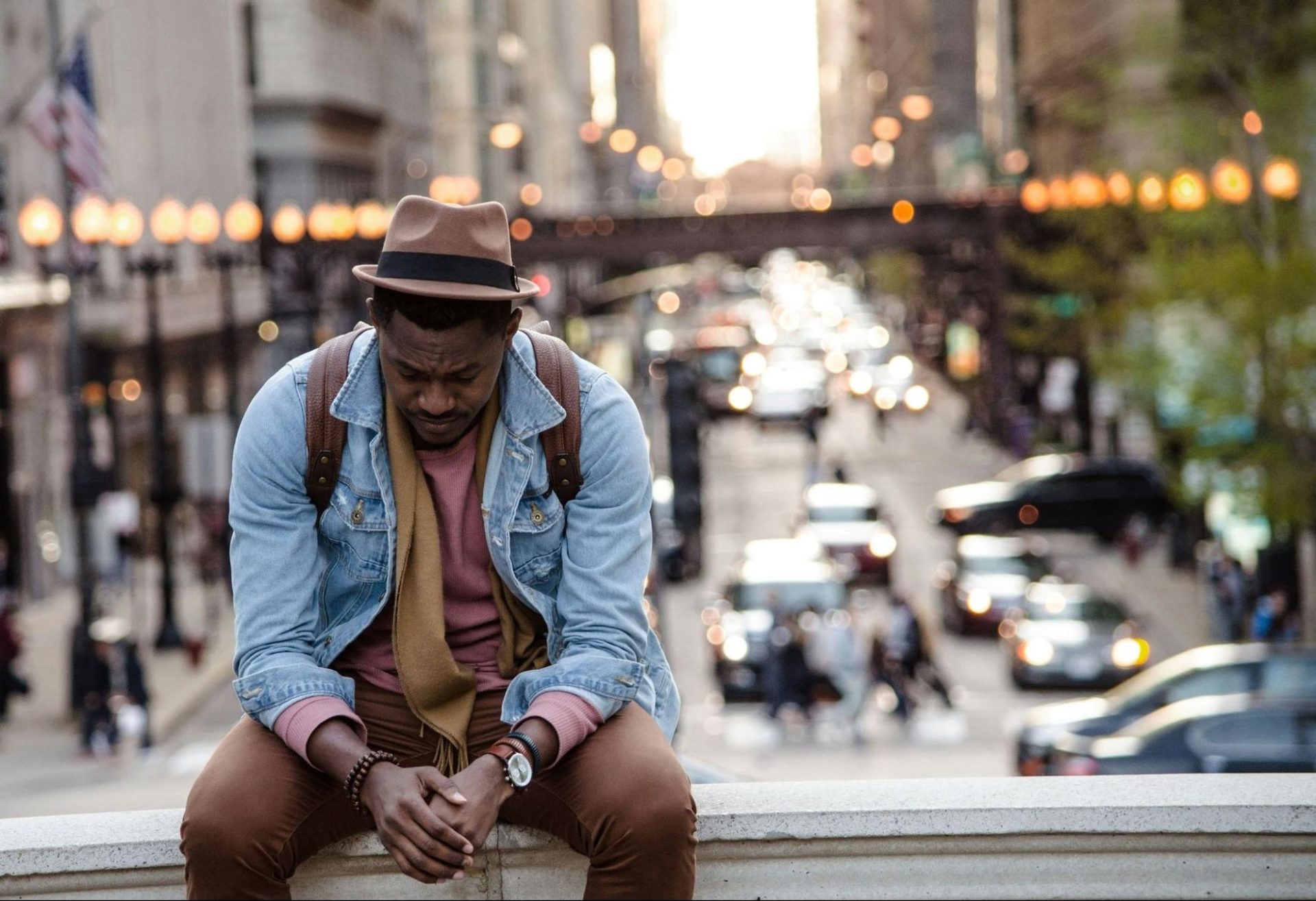 Young man sitting on a concrete ledge in a big city, looking down and worried.