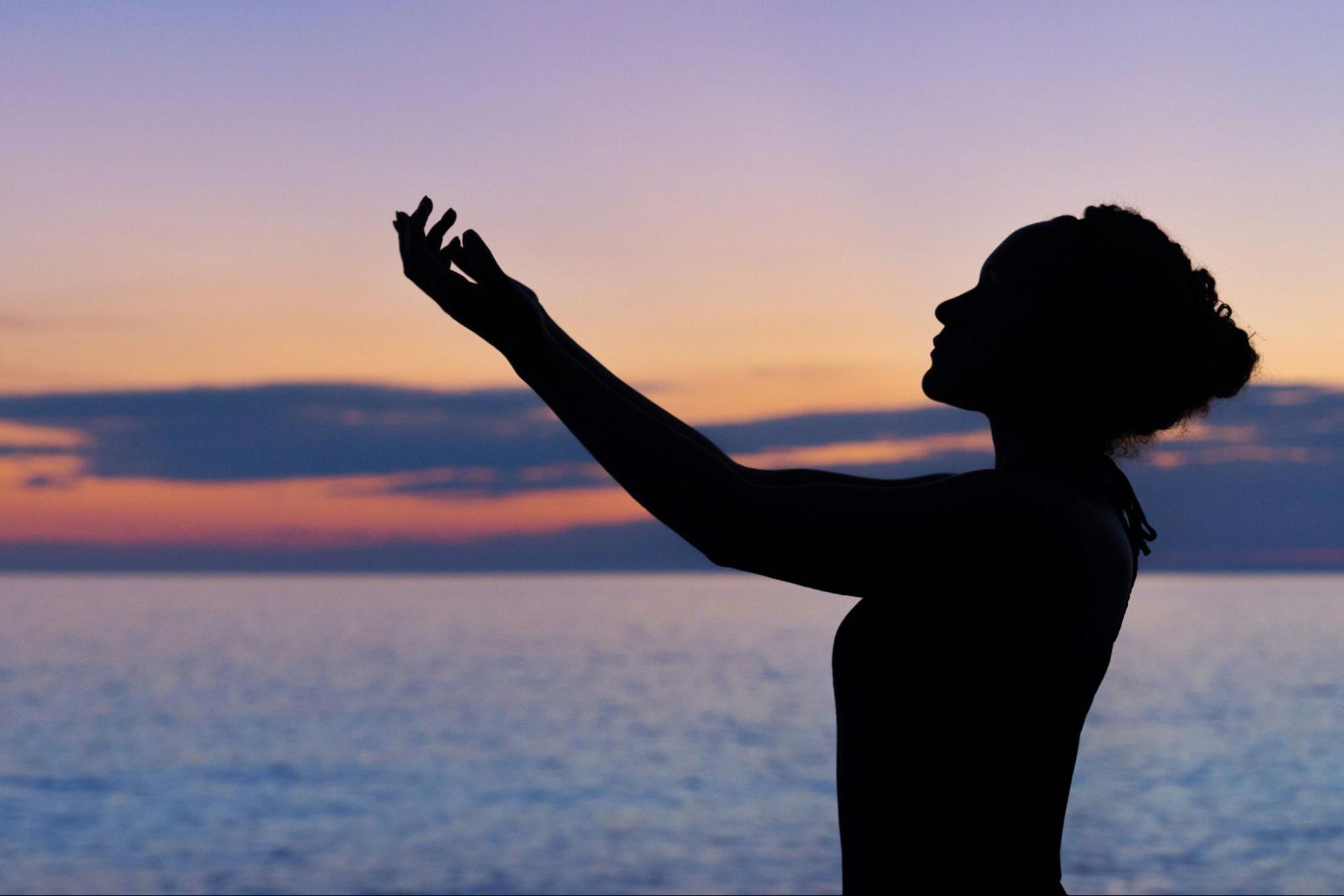 Woman standing in front of ocean at sunset, with hands extended to sky in peace and gratitude. 