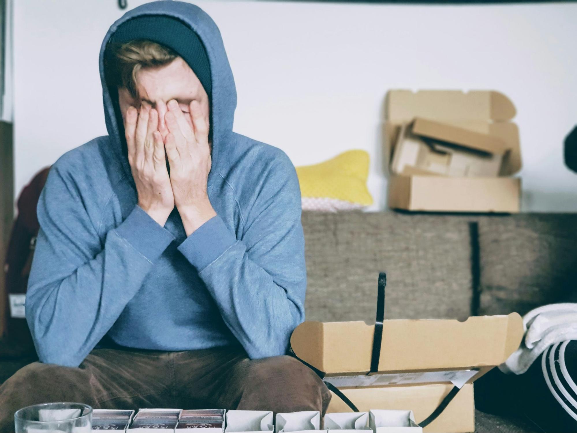 Young man sitting on his counch at home with hands over his face, looking overwhelmed.