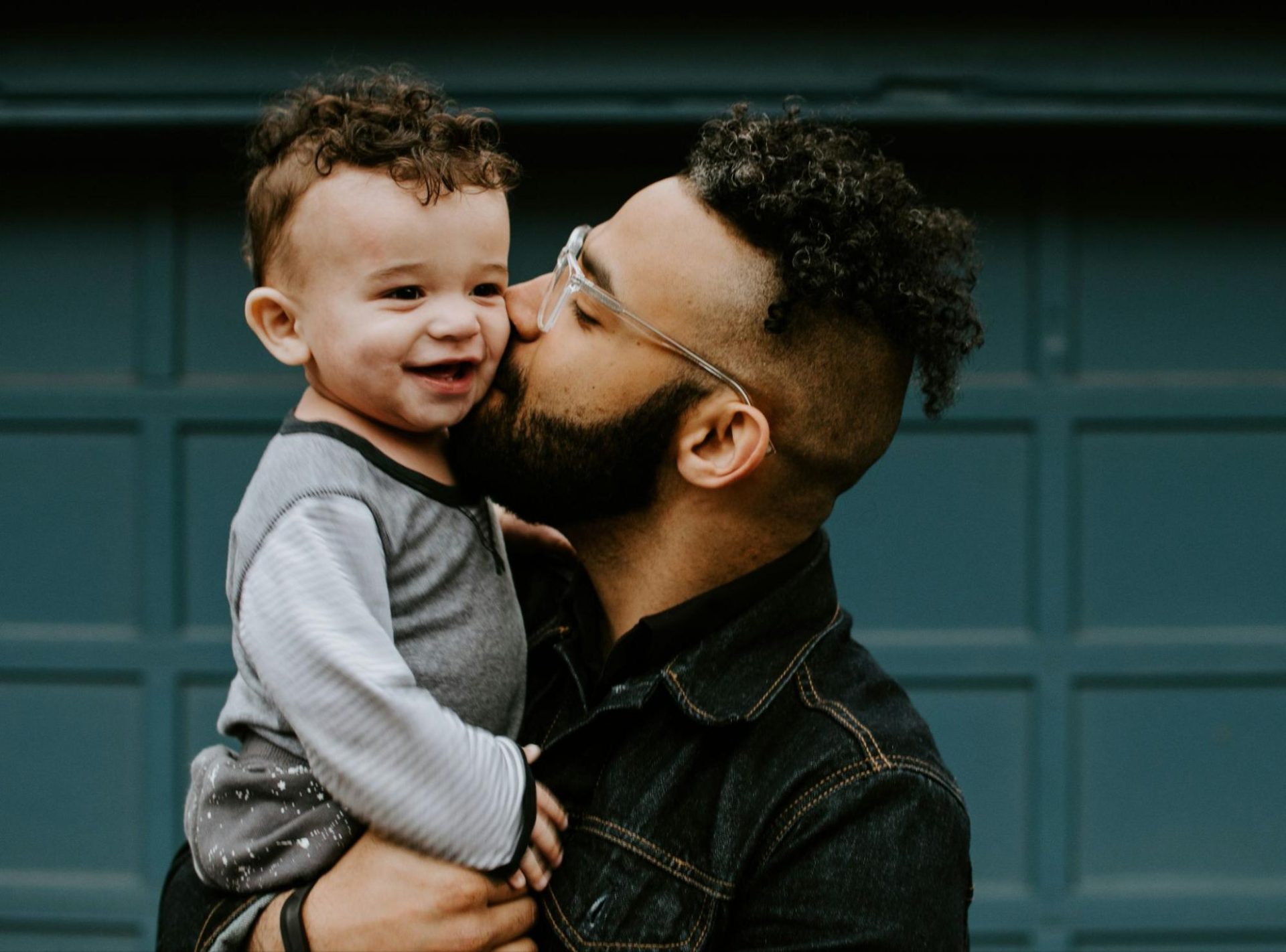 Man with glasses, curly hair and beard holding and kissing 10-month old son.