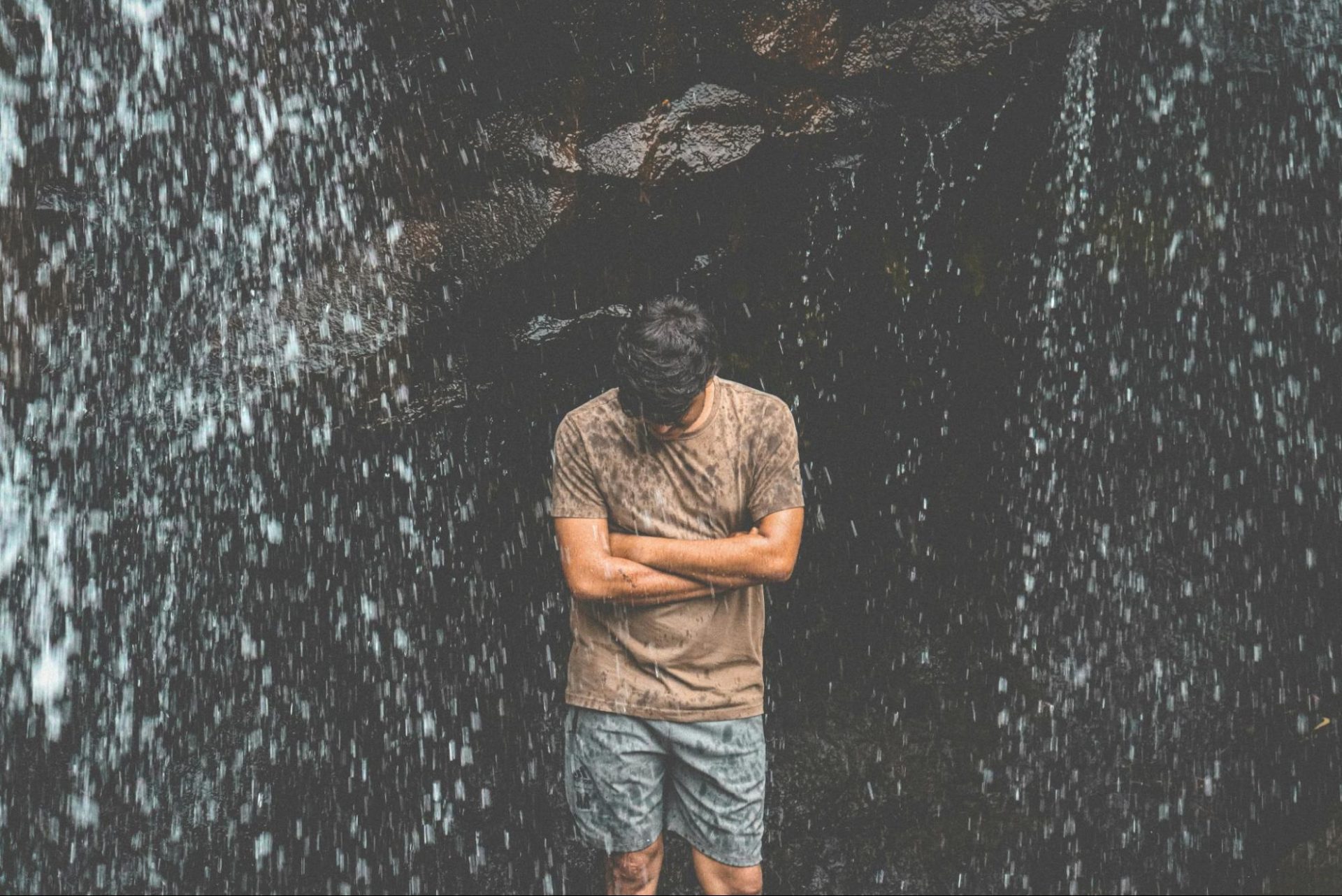 Man standing under a waterfall, arms folded looking down into the ground.