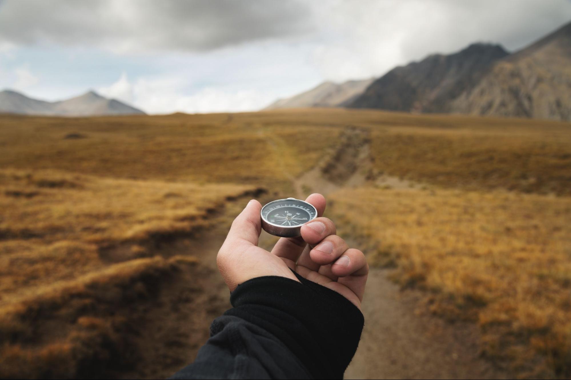 Hand holding a compass in front a long trail through the field surrounded by mountains. 
