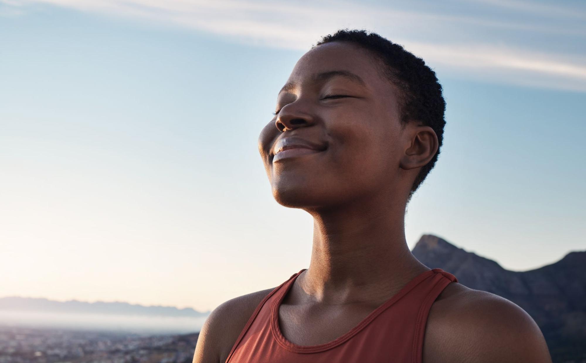 Woman standing in the sun in front of the ocean smiling peacefully with her eyes closed. 